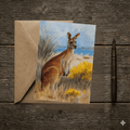 A 5x7 folded greeting card featuring a watercolor painting of an Eastern Grey Kangaroo standing in a field of yellow wattle blossoms and coastal grass, with blue water in the background. The left panel shows a textured off-white finish with the gold ink Willow and Wag Australia logo.