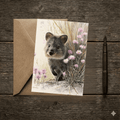 A 5x7 folded greeting card featuring a charming watercolor painting of a smiling quokka lifting its paw among pink daisy wildflowers. The artwork softly fades out into a clean cream-colored background with the gold ink Willow and Wag Australia logo on the back panel.
