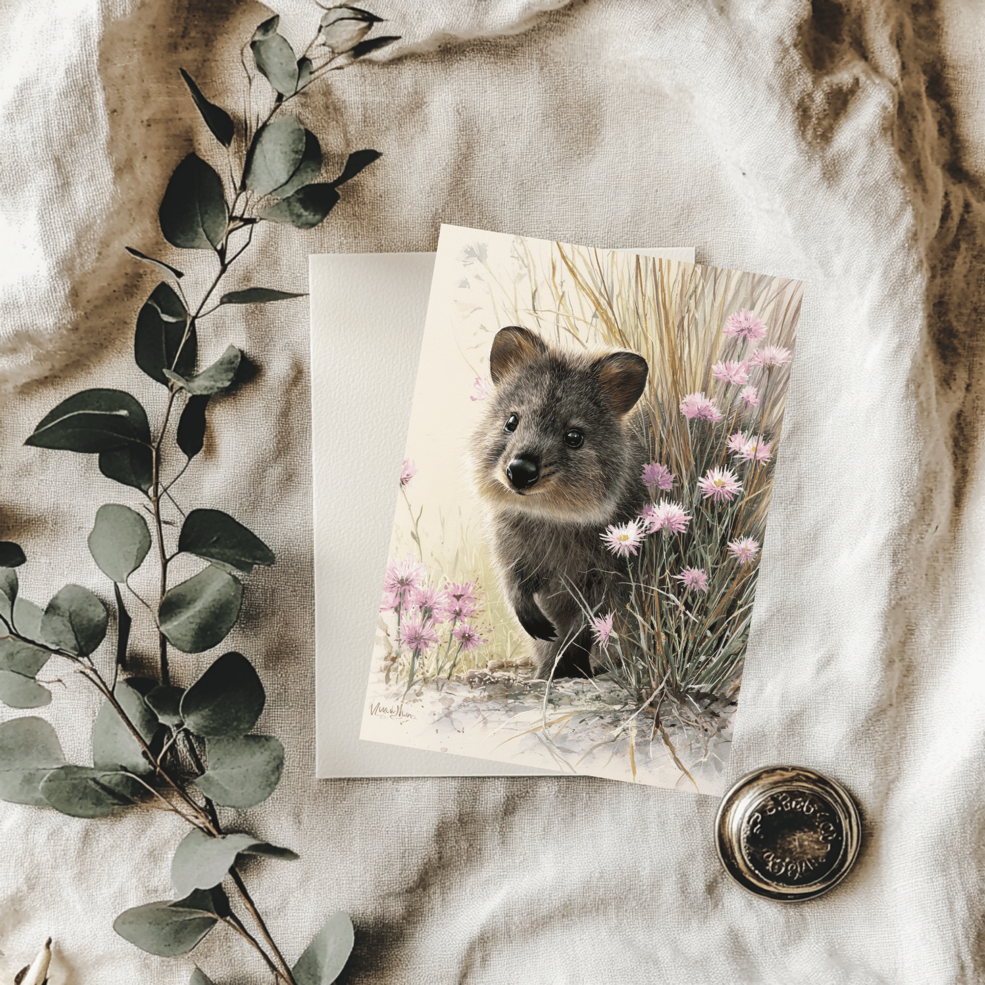A 5x7 folded greeting card featuring a charming watercolor painting of a smiling quokka lifting its paw among pink daisy wildflowers. The artwork softly fades out into a clean cream-colored background with the gold ink Willow and Wag Australia logo on the back panel.