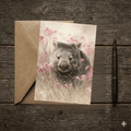 A 5x7 folded greeting card featuring a sweet watercolor painting of a cute wombat looking forward from a field of tall grass and pink heath wildflowers. The left panel shows a textured off-white finish with the gold ink Willow and Wag Australia logo.
